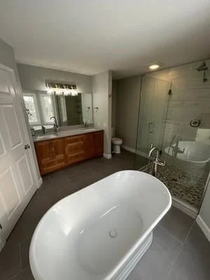 Master bathroom with freestanding white tub, wood double vanity, gray porcelain tile, and frameless glass walk-in shower with pebble floor