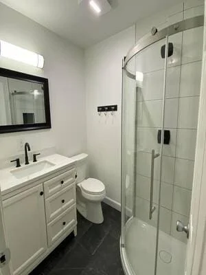 Remodeled bathroom with curved glass shower, gray wall tile, white shaker vanity, black fixtures, and dark gray floor tile