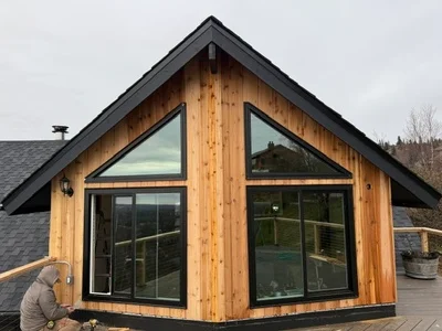 Gabled addition with vertical cedar siding, black-framed sliding glass doors, triangular gable window, and black metal roof trim with a wood deck