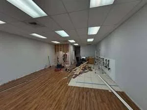Mid-renovation commercial interior with wood-look vinyl plank flooring, removed floor tile, drop ceiling lights, and exposed framing