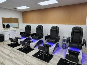 Four black pedicure chairs with blue LED lighting on wood-look plank flooring beside a white vanity and wood-accent wall