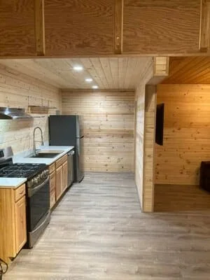 Galley kitchen with knotty pine walls and ceiling, wood cabinets, black gas range, stainless fridge, and gray vinyl plank floor