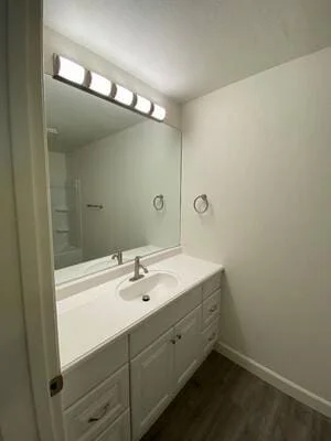 White vanity with integrated sink, chrome faucet, three-bulb light bar above large mirror, dark vinyl plank flooring
