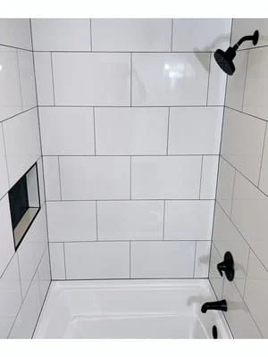 White large-format wall tile with black grout around a white tub, matte black shower head and tub spout, and a recessed tile niche