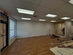 Commercial room mid-demolition with drop ceiling, fluorescent troffer lights, bare drywall, and wood-look vinyl flooring with debris
