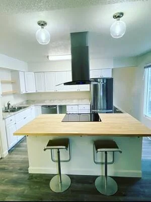 Kitchen with butcher block island, black cooktop, stainless range hood, white cabinets, and dark wood-look flooring