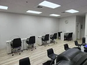 Commercial nail salon interior with gray vinyl plank flooring, LED ceiling lights, and white manicure tables with black chairs