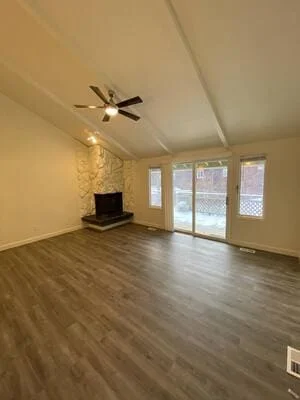 Empty living room with gray-brown luxury vinyl plank flooring, stone fireplace, vaulted ceiling with exposed beams, and sliding glass doors